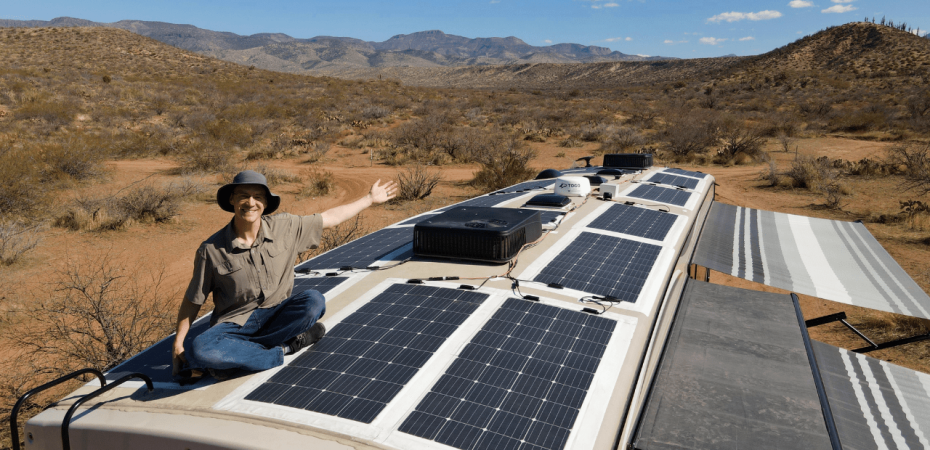 Man showcasing solar panels on RV roof in desert landscape.